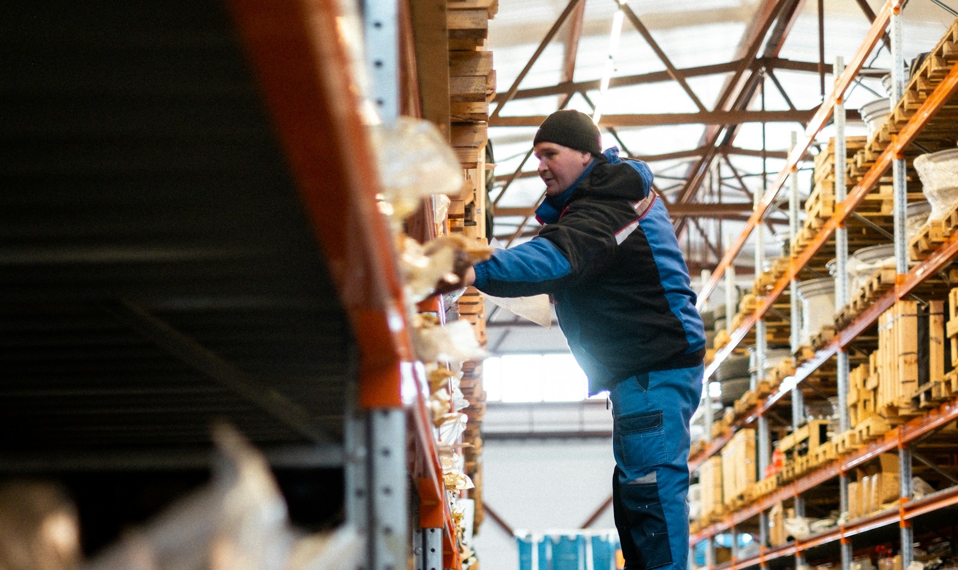 A warehouse worker in a blue and black uniform reaching for items on a high storage shelf.